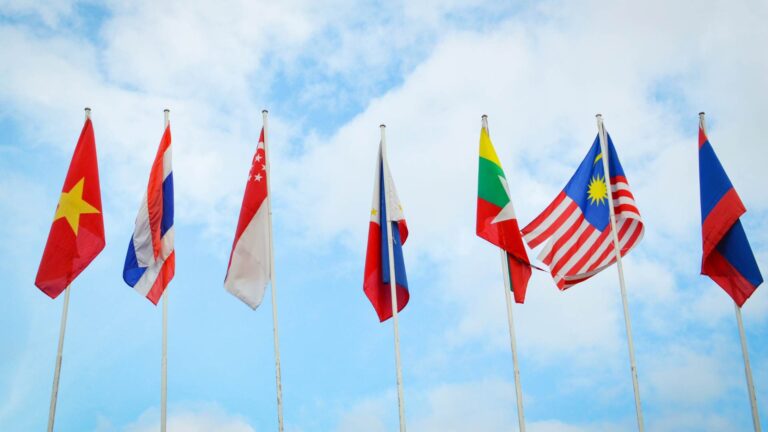 National flags of several countries, including Vietnam, Thailand, Singapore, the Philippines, Myanmar, Malaysia, and Cambodia, displayed on tall flagpoles against a blue sky with light clouds, representing international trade and global markets.