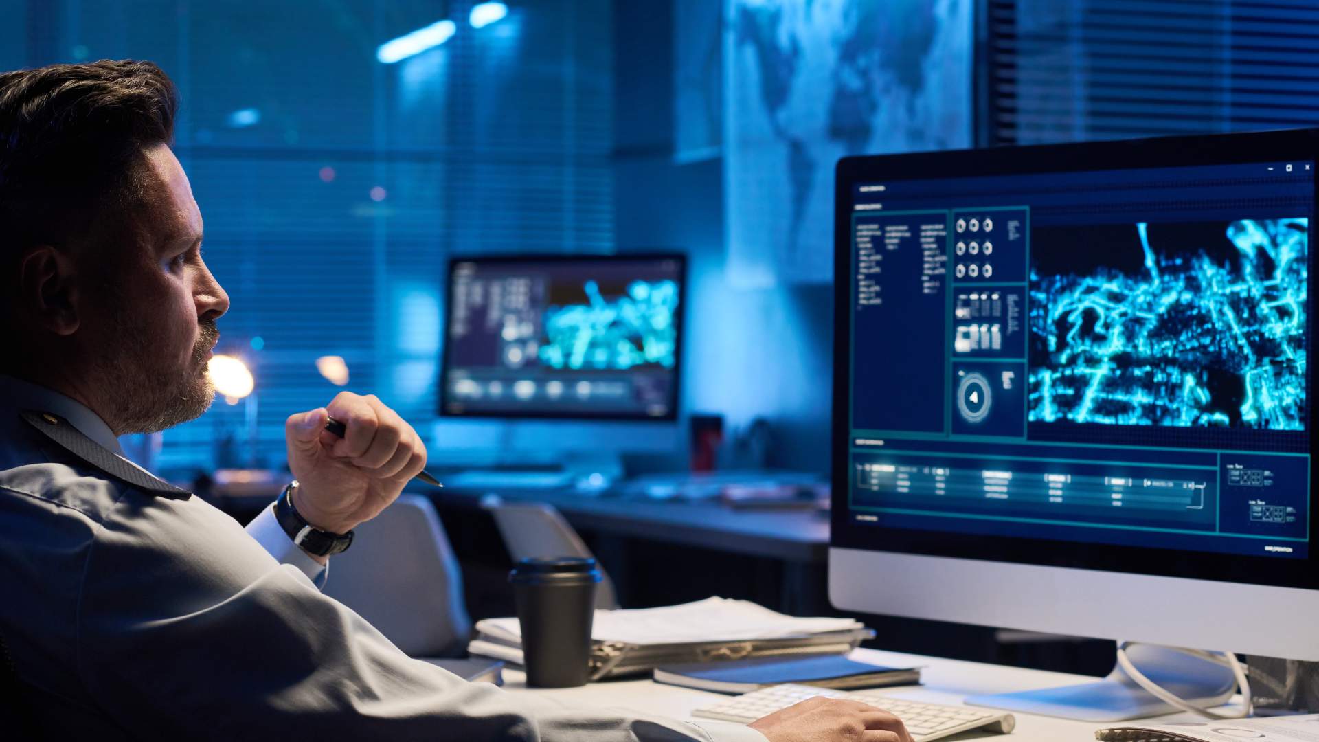 Professional man in an office at night analyzing data on a large desktop monitor displaying blue-toned digital dashboards and network visuals.