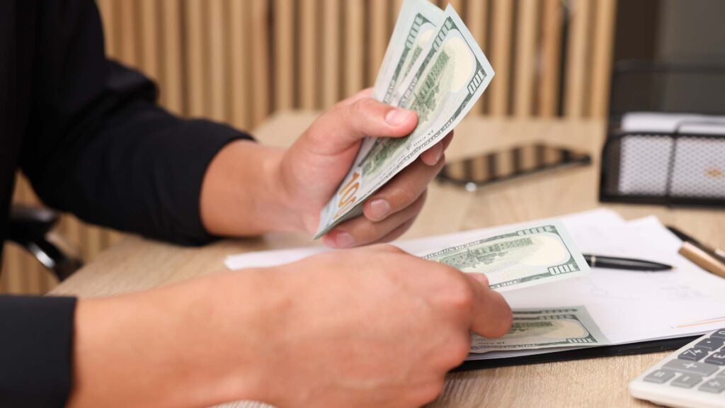 Close-up of a person counting U.S. dollar bills at a desk with documents, calculator, and pen, representing financial transactions or cost calculations.