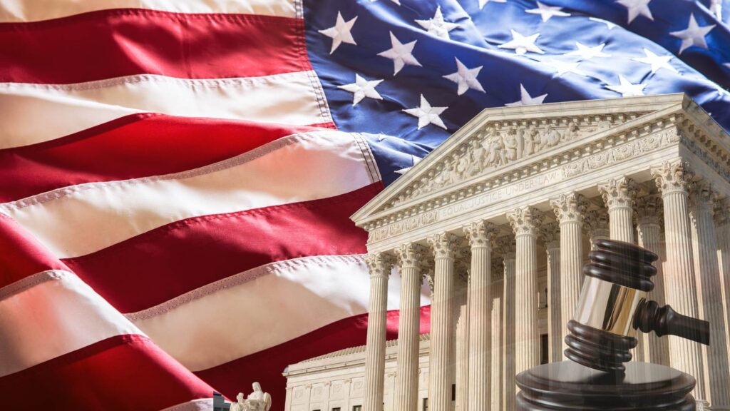 Close-up of the U.S. Supreme Court building with its columns and pediment in front of a large waving American flag, alongside a wooden judge’s gavel, symbolizing a major Supreme Court ruling on U.S. trade and tariff authority.