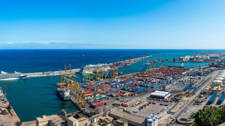 Aerial view of a busy container port with cargo ships, stacked shipping containers, and large cranes loading freight along a coastal harbor.