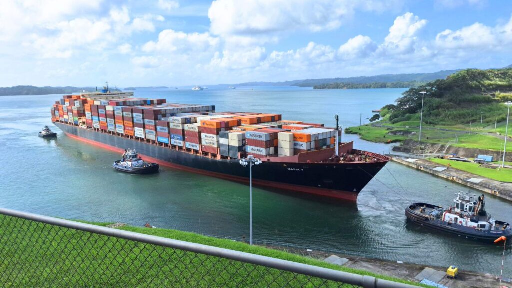 Container ship loaded with stacked cargo containers transiting a canal with tugboats assisting, representing global trade and international shipping.