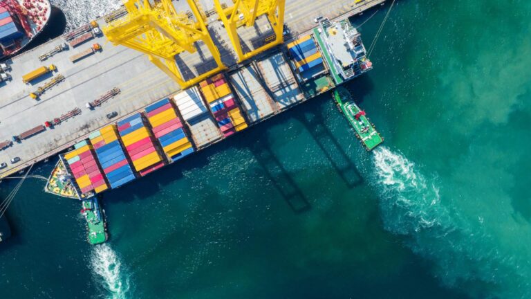 Container ship at port with cranes loading cargo and tugboats assisting, representing global trade and import/export logistics